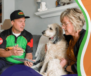 A ClearView Services technician in a green and black uniform sits on a couch with a tablet, speaking to a woman holding her dog. The technician is explaining services in a friendly, professional manner inside a cozy living room.