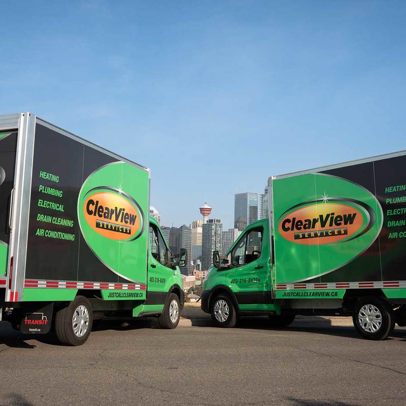 A ClearView Services technician in a green and black uniform sits on a couch with a tablet, speaking to a woman holding her dog. The technician is explaining services in a friendly, professional manner inside a cozy living room.